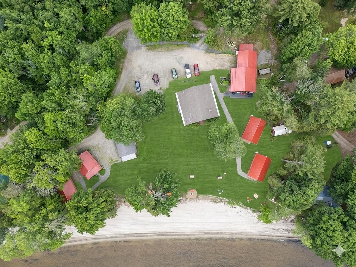 Overhead aerial of the two-acre compound, six cabin rooflines tucked among pines, the central lodge centered on a wide green clearing, sand beach below.