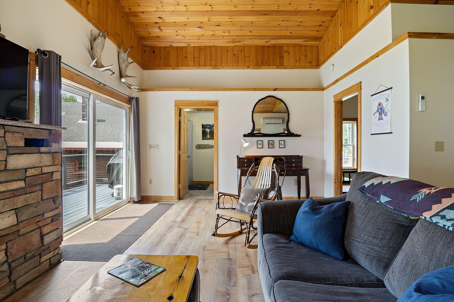 Cabin interior with pine-paneled ceiling, stone fireplace, antler wall mount, and a lakeside slider door.
