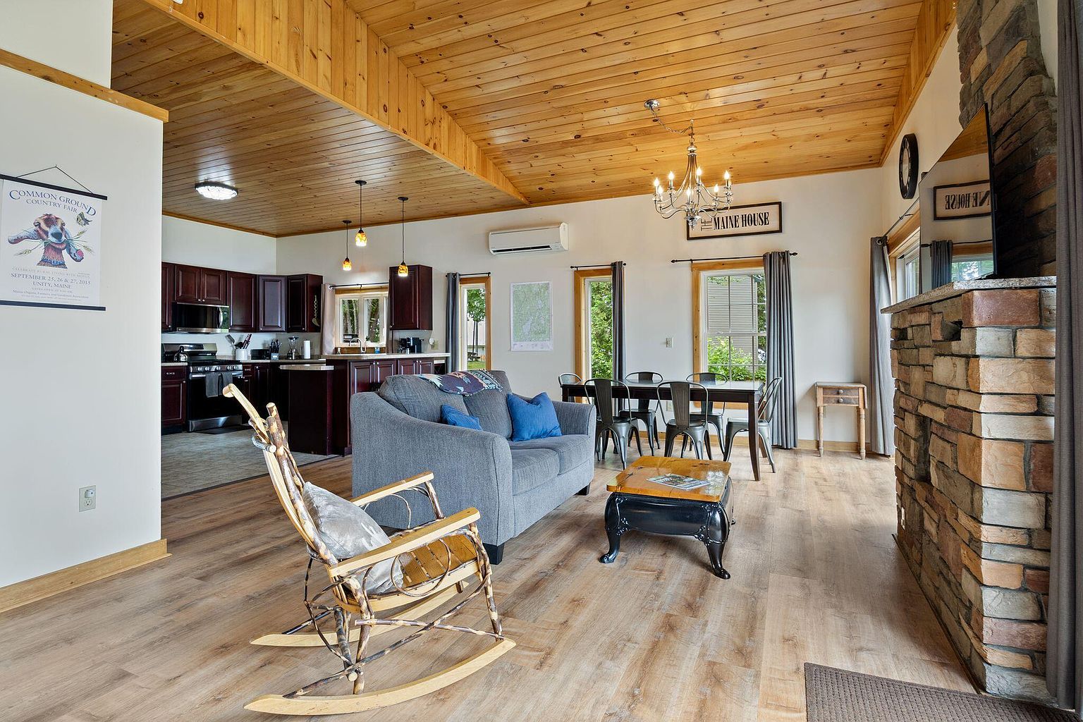 Open great room of a renovated cabin with vaulted pine ceiling, dark wood kitchen, dining table for eight, and a hand-built log rocker.
