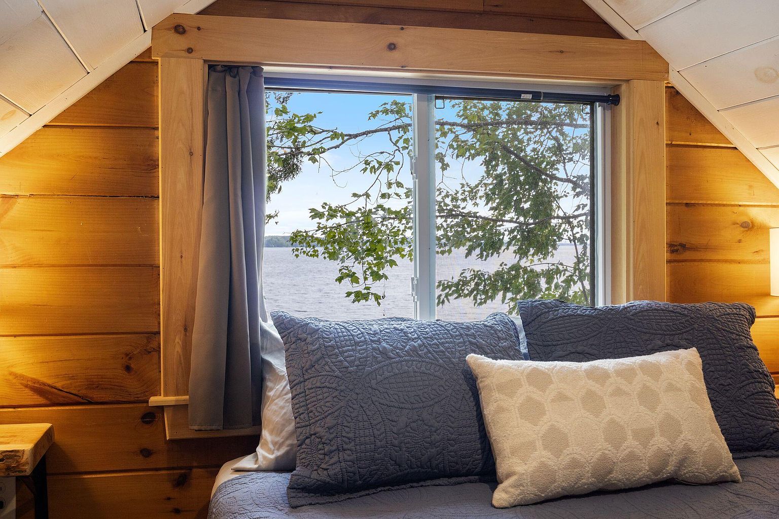 A cabin loft bed framed by a small window looking out over Lake Winnecook, branches in soft focus.