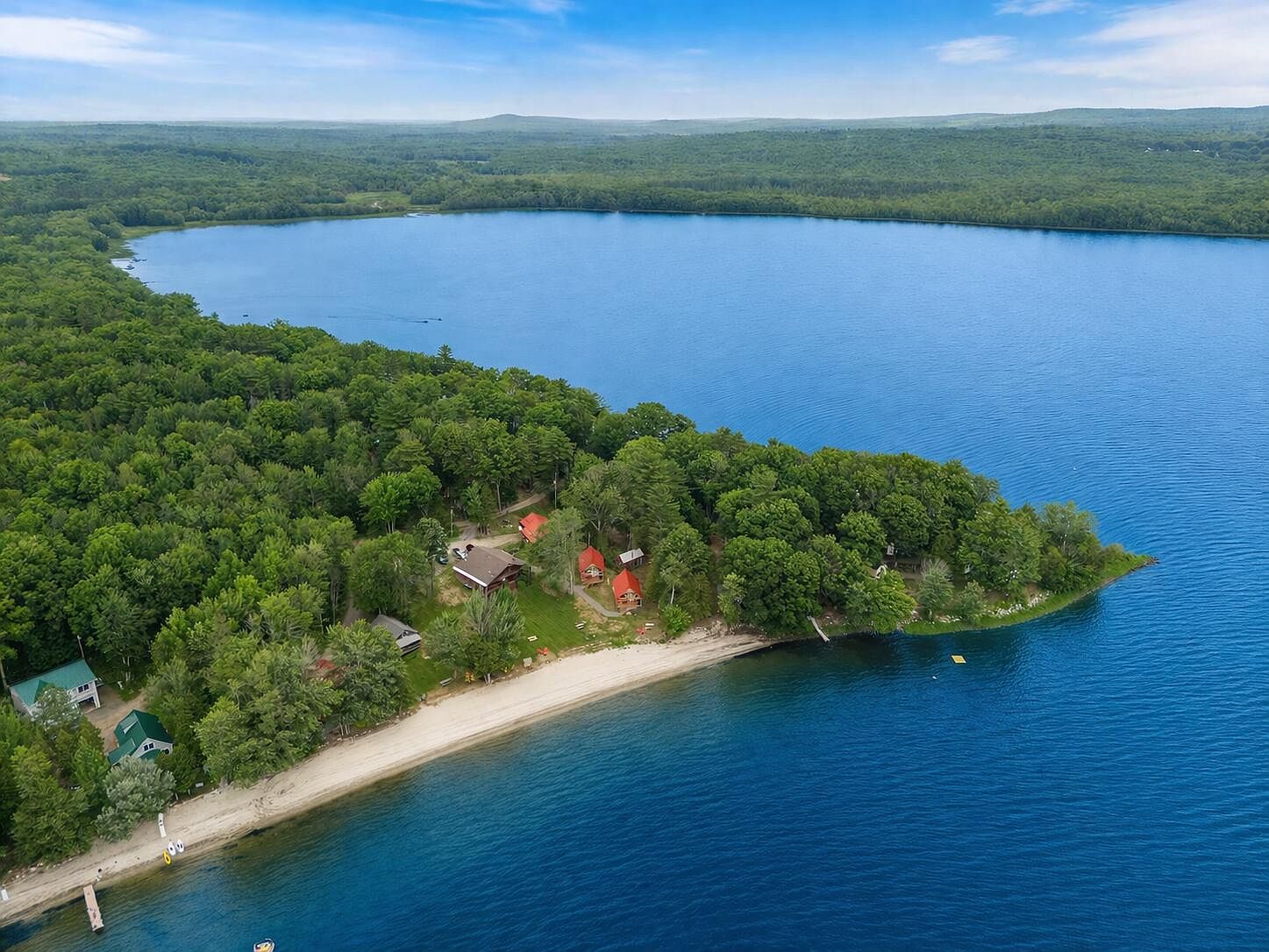 Aerial view of The Lodge and Cabins at Lake Winnecook, showing the two-acre compound, a private sandy beach curving into deep blue water, six cabins set among the pines, and the far Maine shoreline.